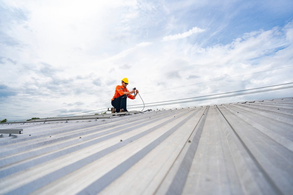 Technician in safety uniform and helmet kneeling on a rooftop, using a drill for precise installation work
