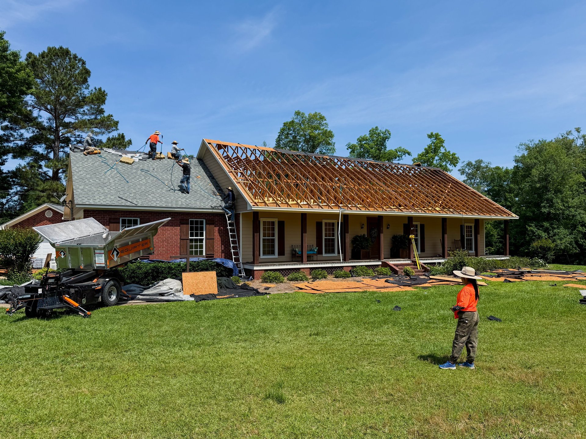House under renovation with exposed wooden framing, workers on roof, construction materials in yard, surrounded by green lawn and trees
