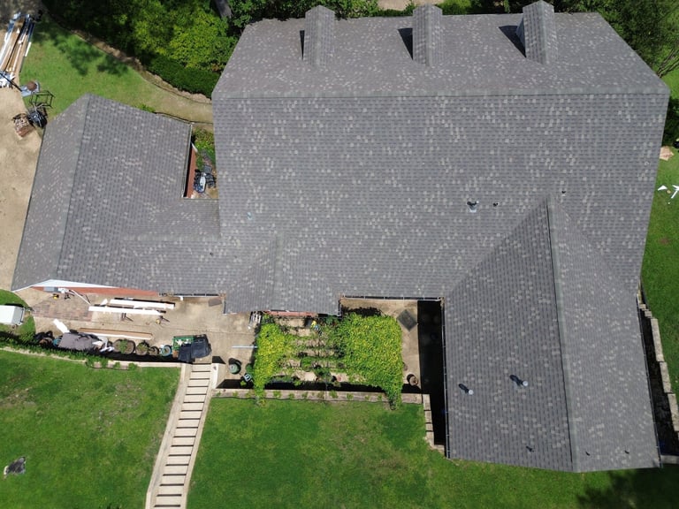 Aerial view of a residential house with gray shingle roof, connected garage, and manicured yard with green lawn and landscaping.