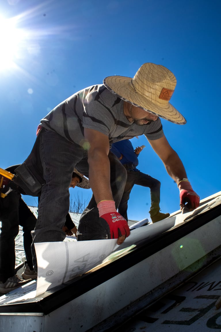 Worker in straw hat and safety gear installing white pipe on roof under bright blue sky