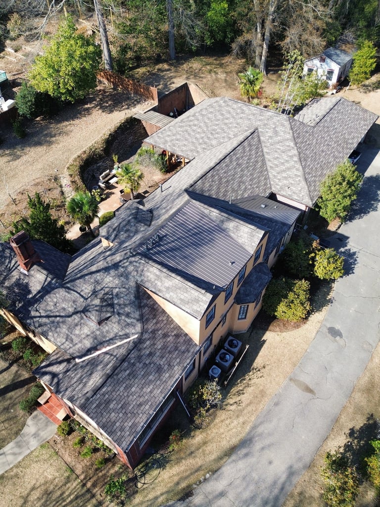 Aerial view of a residential home with gray shingle roof, patio cover, and surrounding landscape with trees