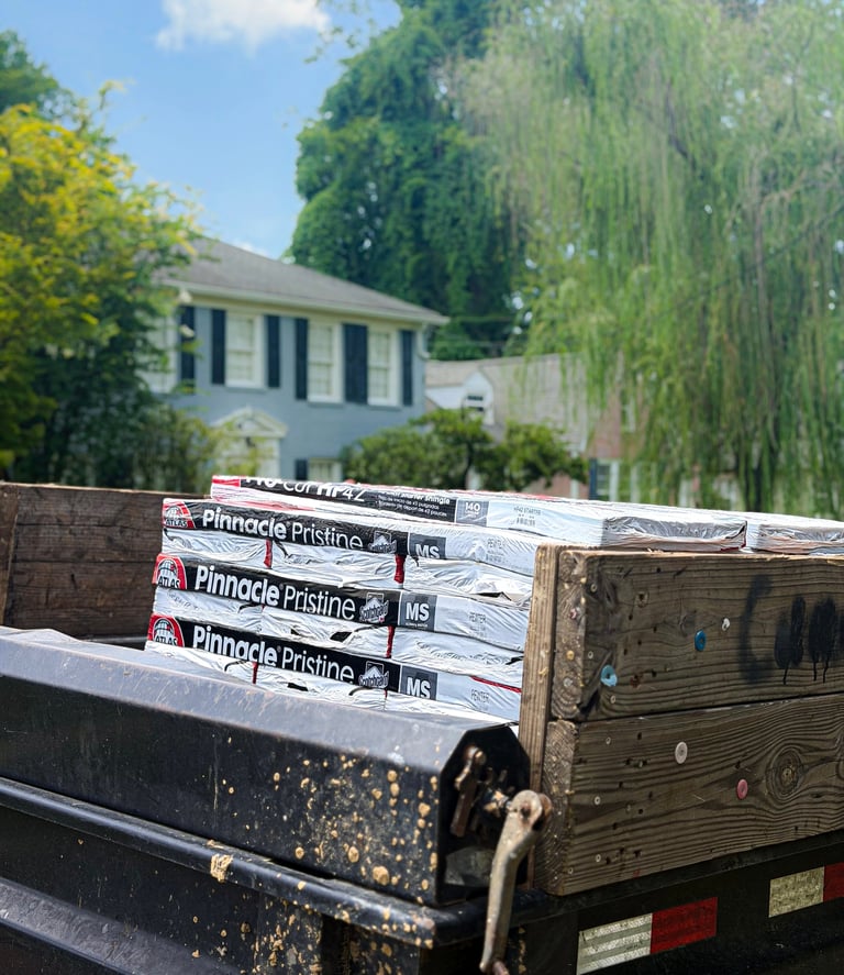 Stacked rolls of Pinnacle Pristine roofing material on a wooden truck bed with a residential house and trees in background