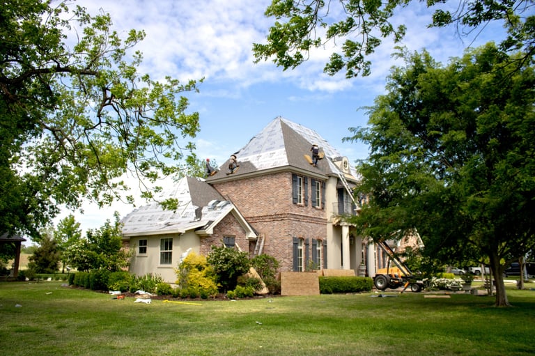 Brick Victorian house with mansard roof surrounded by green lawn and mature trees on a sunny day