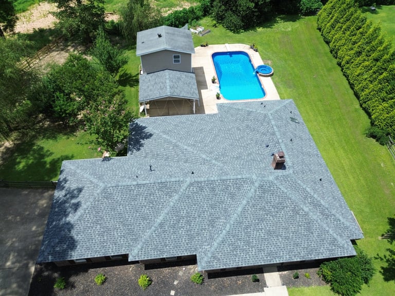 Aerial view of a residential home with gray roof, pool deck, and blue rectangular swimming pool surrounded by green lawn and trees