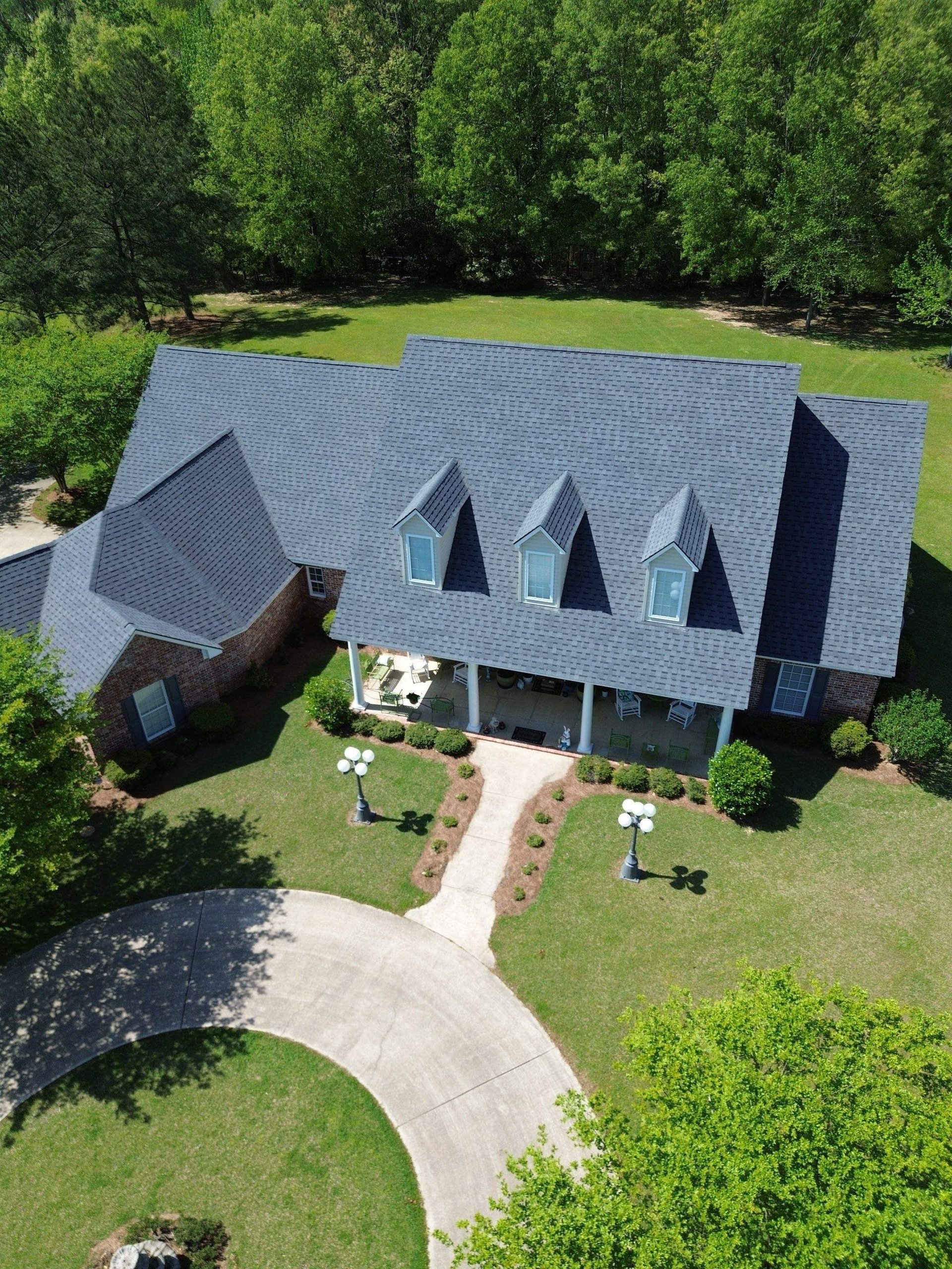 Aerial view of a modern two-story home with gray shingle roof and arched windows, surrounded by green lawn and trees