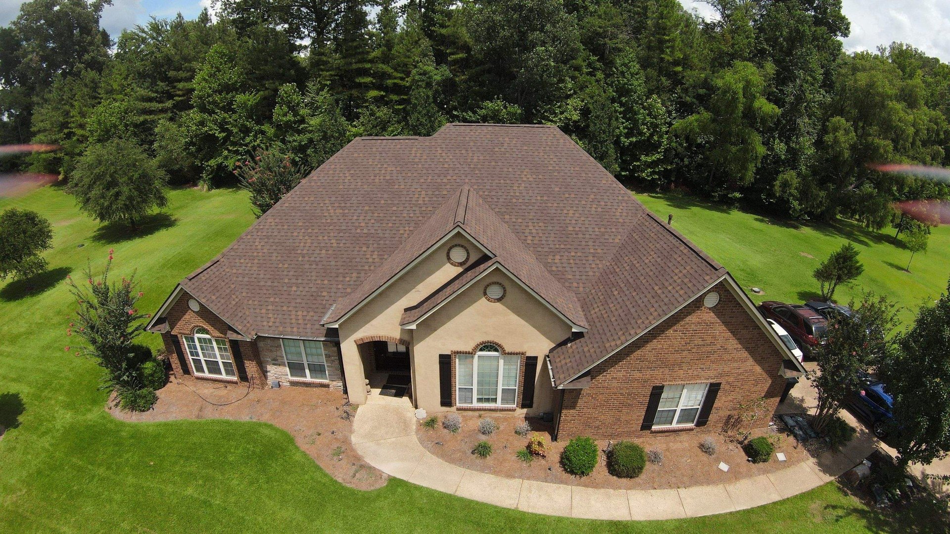 Aerial view of a brick single-family home with brown roof surrounded by green lawn and mature trees