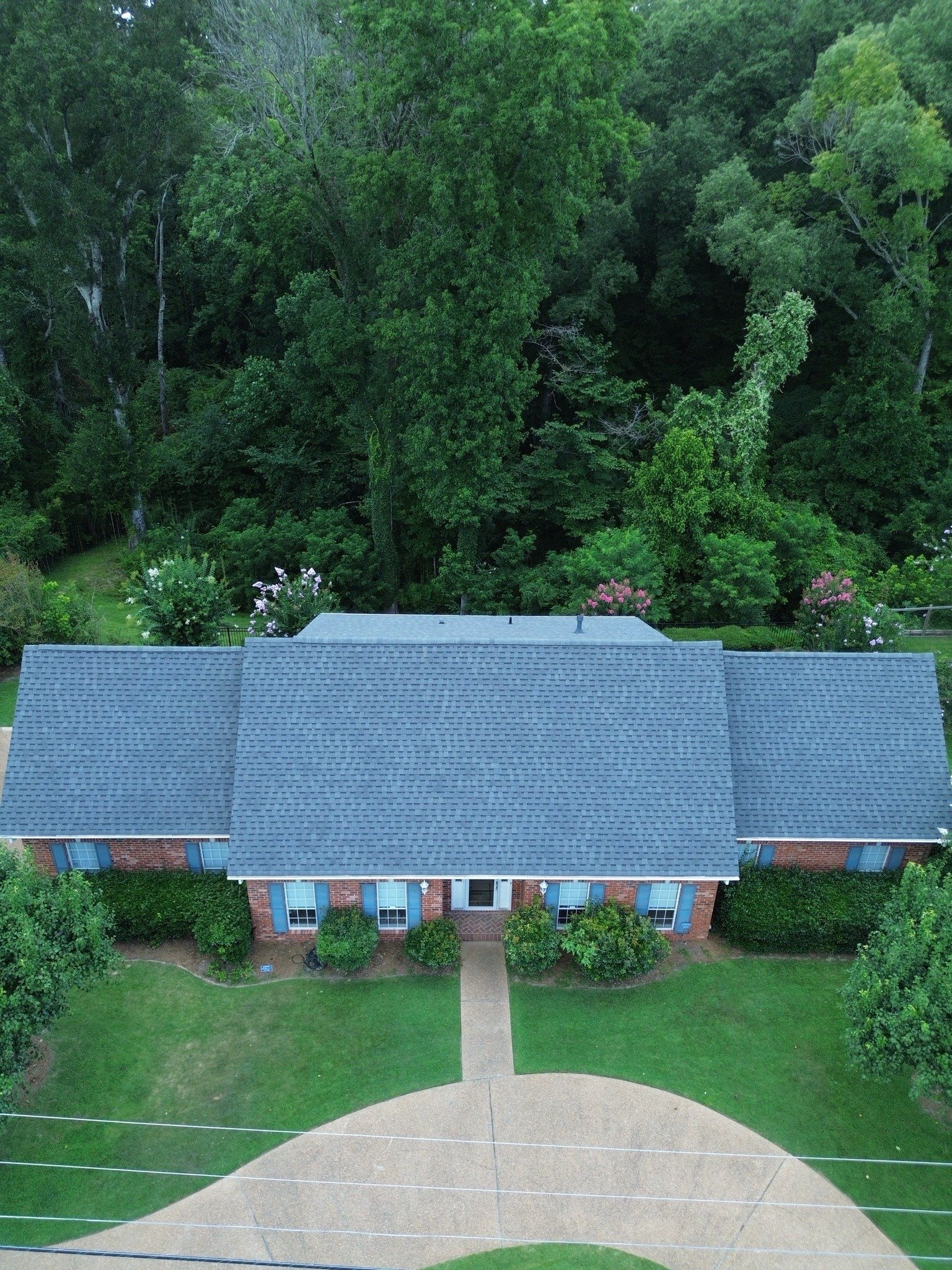 Aerial view of a brick house with blue-gray roof surrounded by green lawn and dense trees
