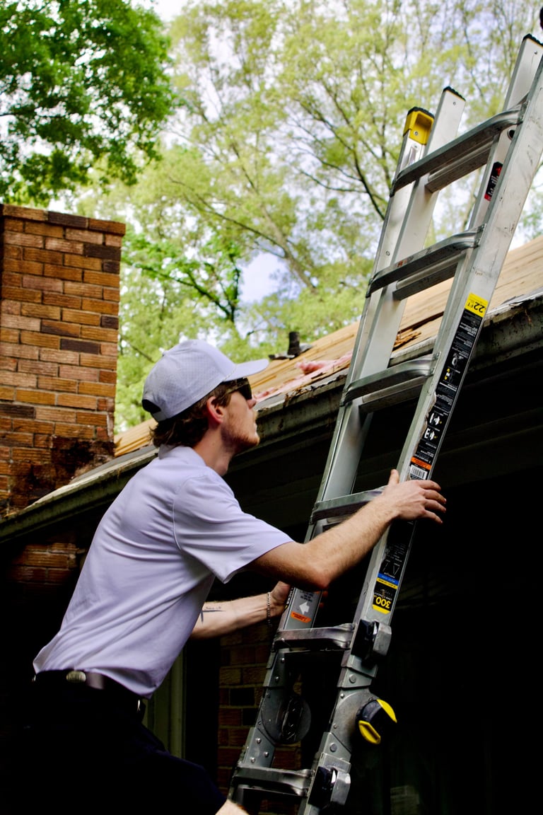 Worker in cap and light shirt positioning aluminum ladder against brick building with roof and trees in background