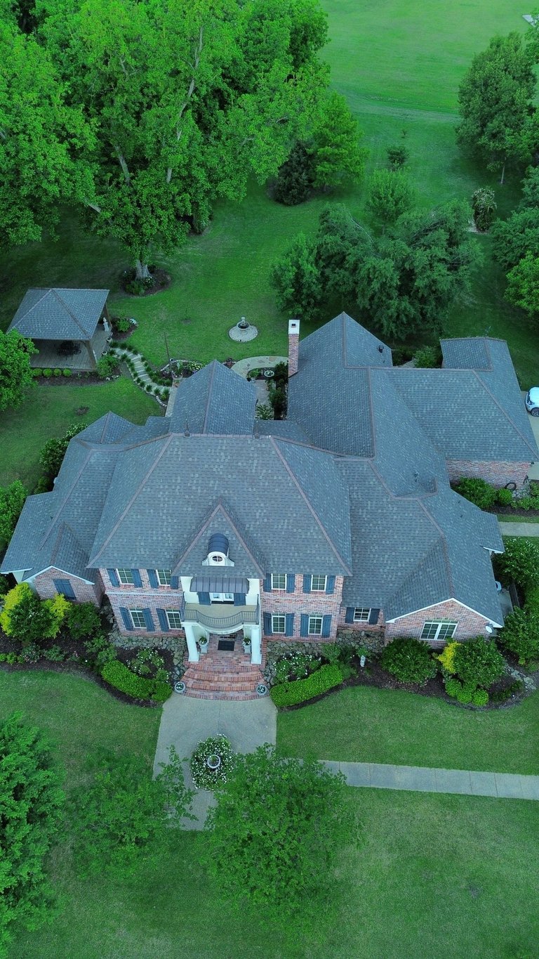 Aerial view of a large brick mansion with slate gray roofing surrounded by manicured green lawn and mature trees.