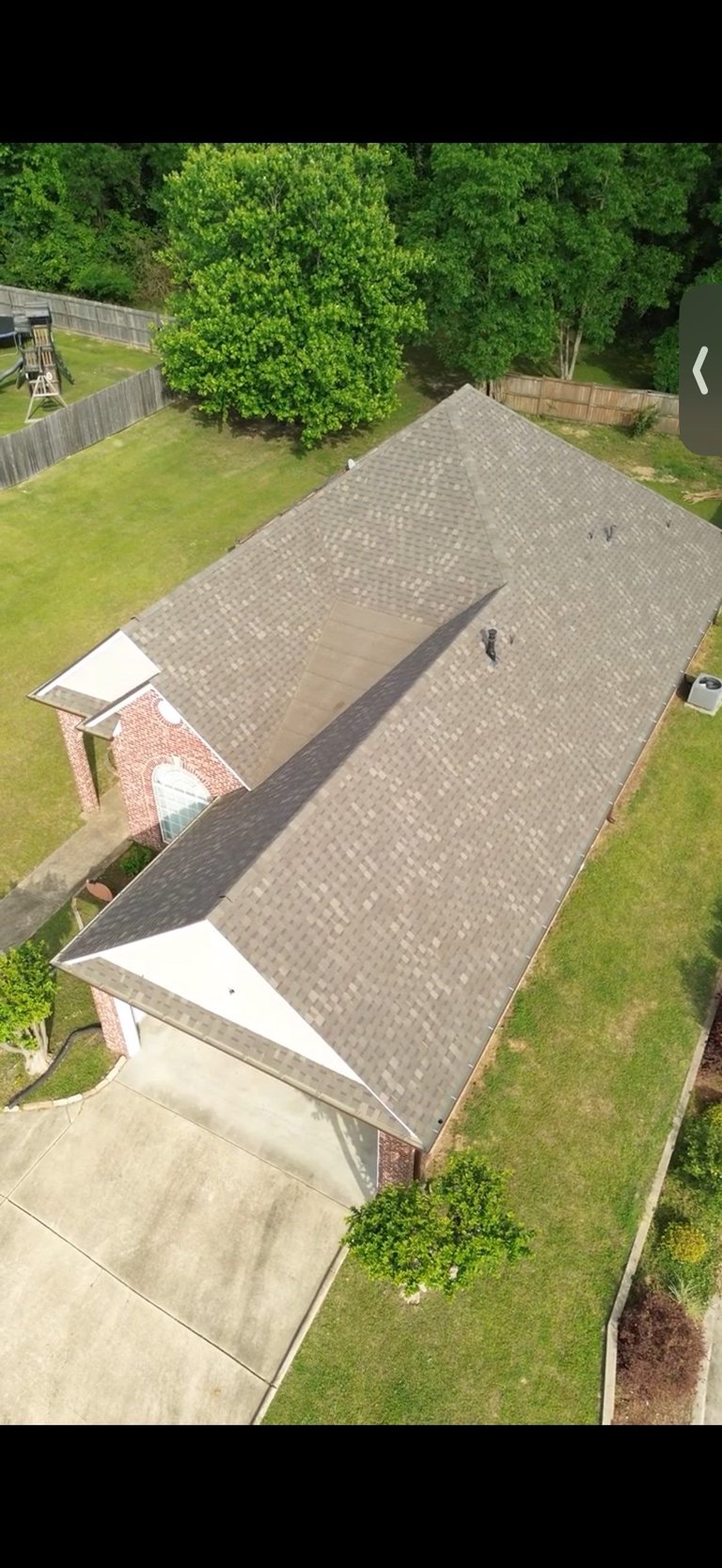 Aerial view of a residential house with gray shingle roof, concrete driveway, and green lawn surrounded by trees and fenced yard