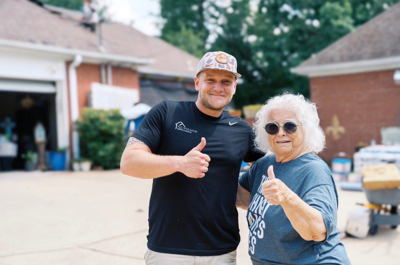 Man in black shirt and cap gives thumbs up next to elderly woman in sunglasses in a driveway