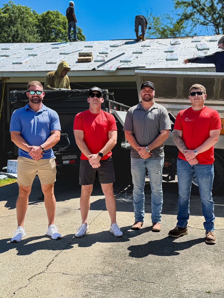 Four workers in casual attire posing in front of a house with roof installation in progress and workers on top