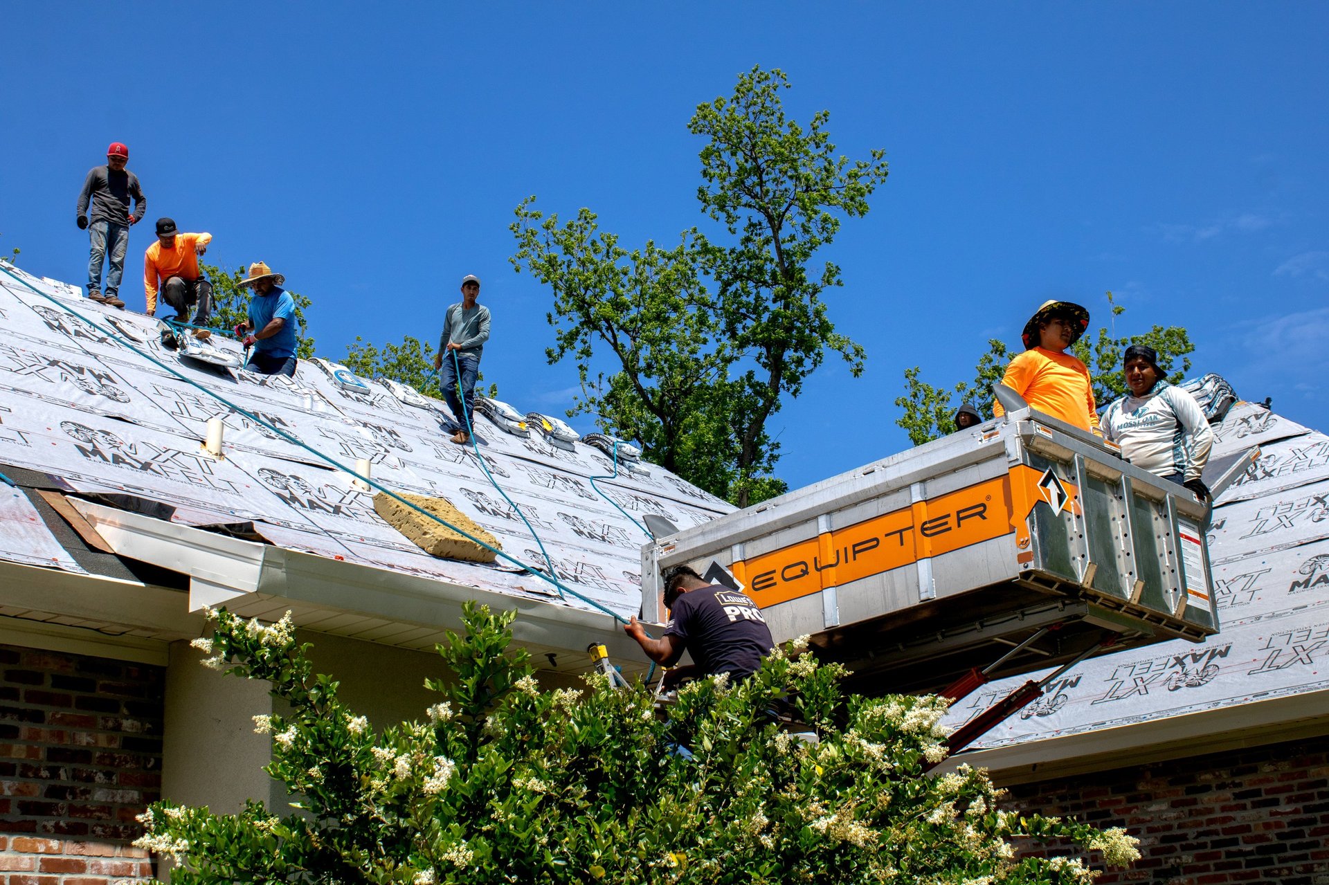 Construction workers installing white roof material on a residential home with a yellow dump truck, clear blue sky visible above