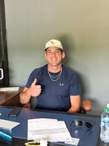 Man in navy Under Armour shirt and white baseball cap giving thumbs up at desk with documents and water bottle