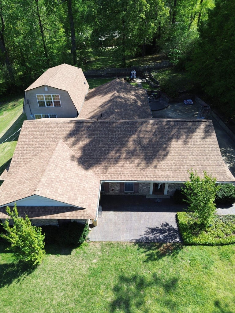 Aerial view of a residential house with brown roof, driveway, garage, and surrounding green trees and lawn
