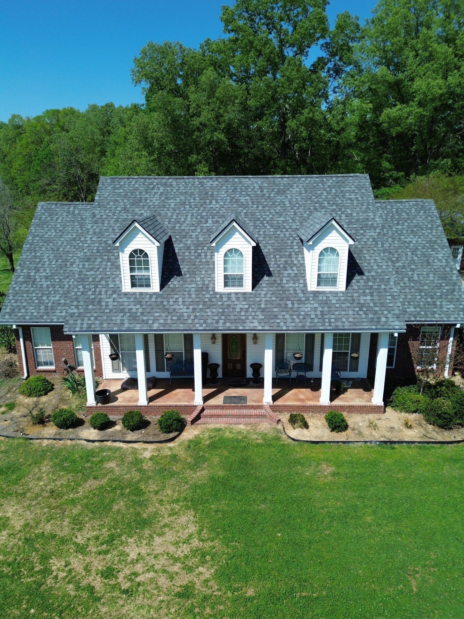 Farmhouse with gray roof, white columns, three dormers, brick porch, and manicured lawn surrounded by trees