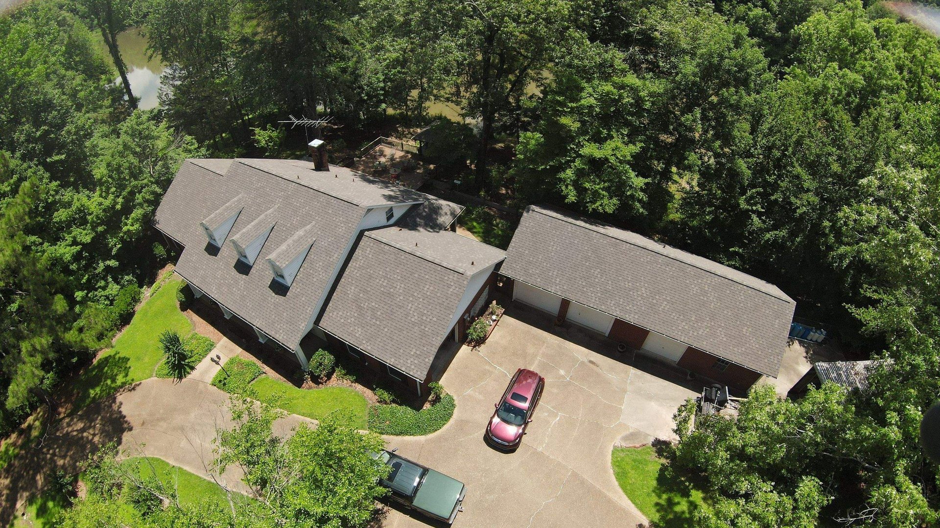 Aerial view of a brick house with blue-gray roof surrounded by green lawn and dense trees